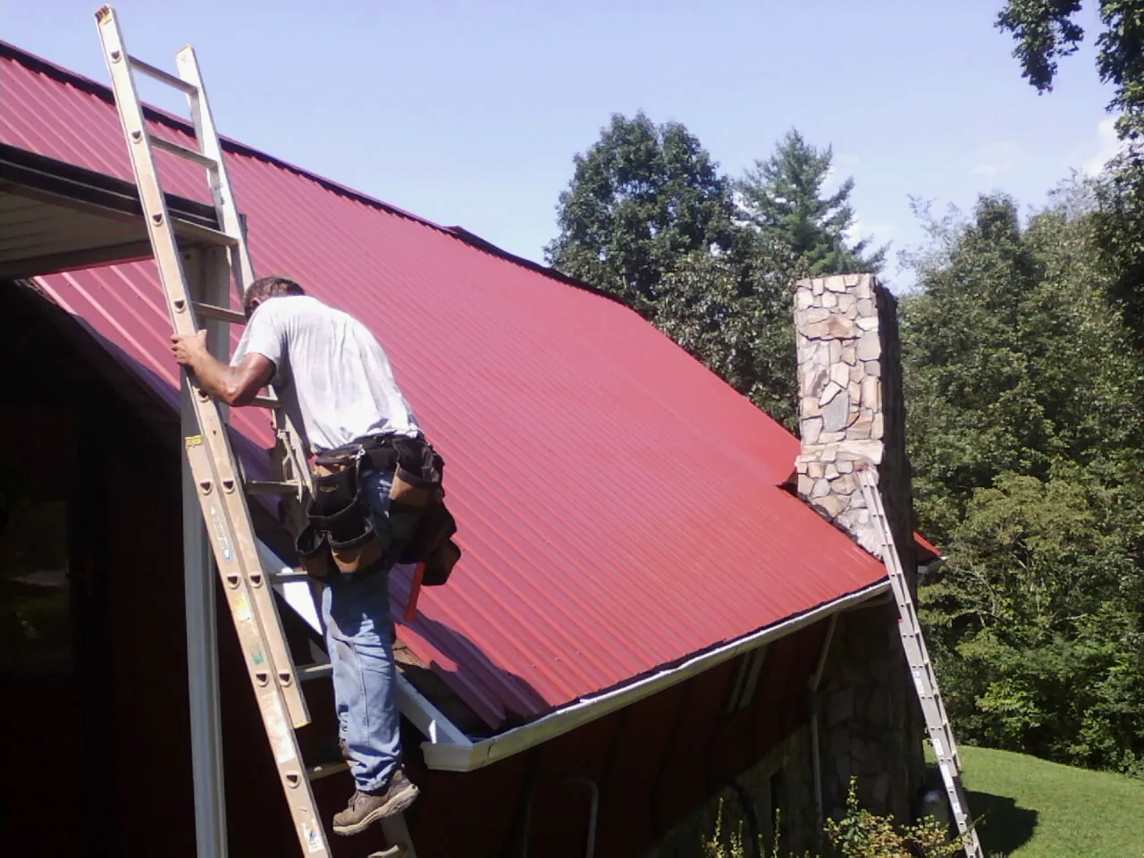 Roofer on a ladder beside a red metal roof and stone chimney — Soffit & Fascia Repair by Roofing Brothers in Bailey Island